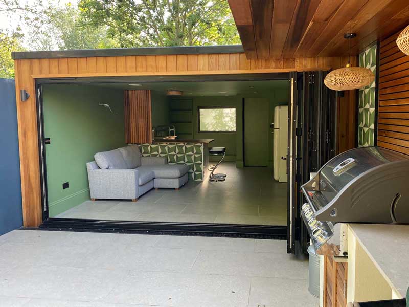 timber clad garden room from Aoba Gardens with comfy sofas within and an outdoor kitchen just outside