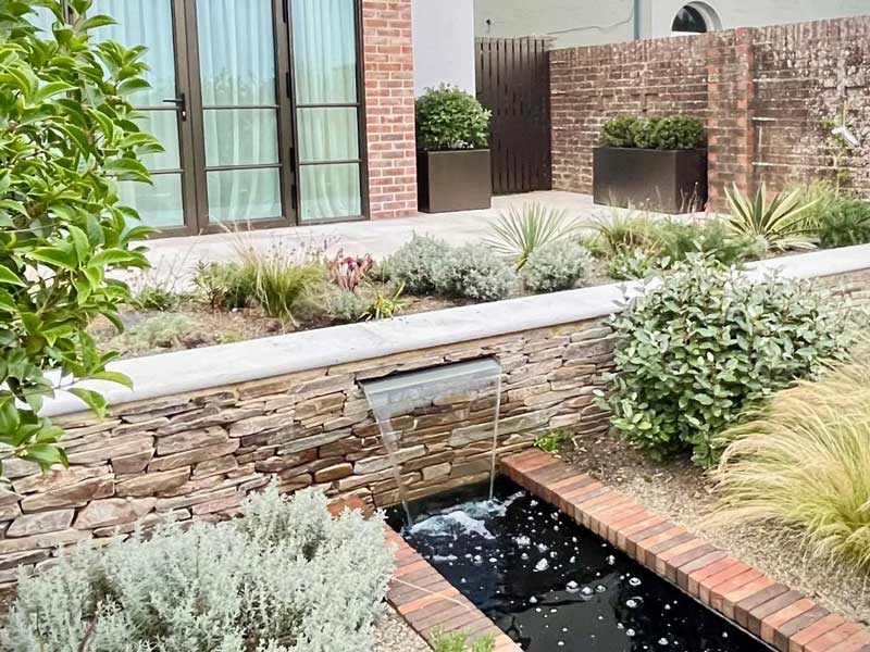 A water blade set into a paddlestone retaining wall, pours clear water into a long rill that runs through a beautifully planted border