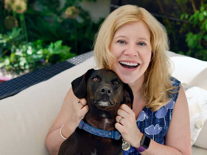 Head and shoulders image of garden designer Judy Shardlow wearing a blue top and cuddling a small black dog