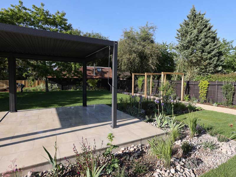 lawned garden bisected by a raised patio shaded by a modern pergola and surrounded by gravel-mulched planting beds